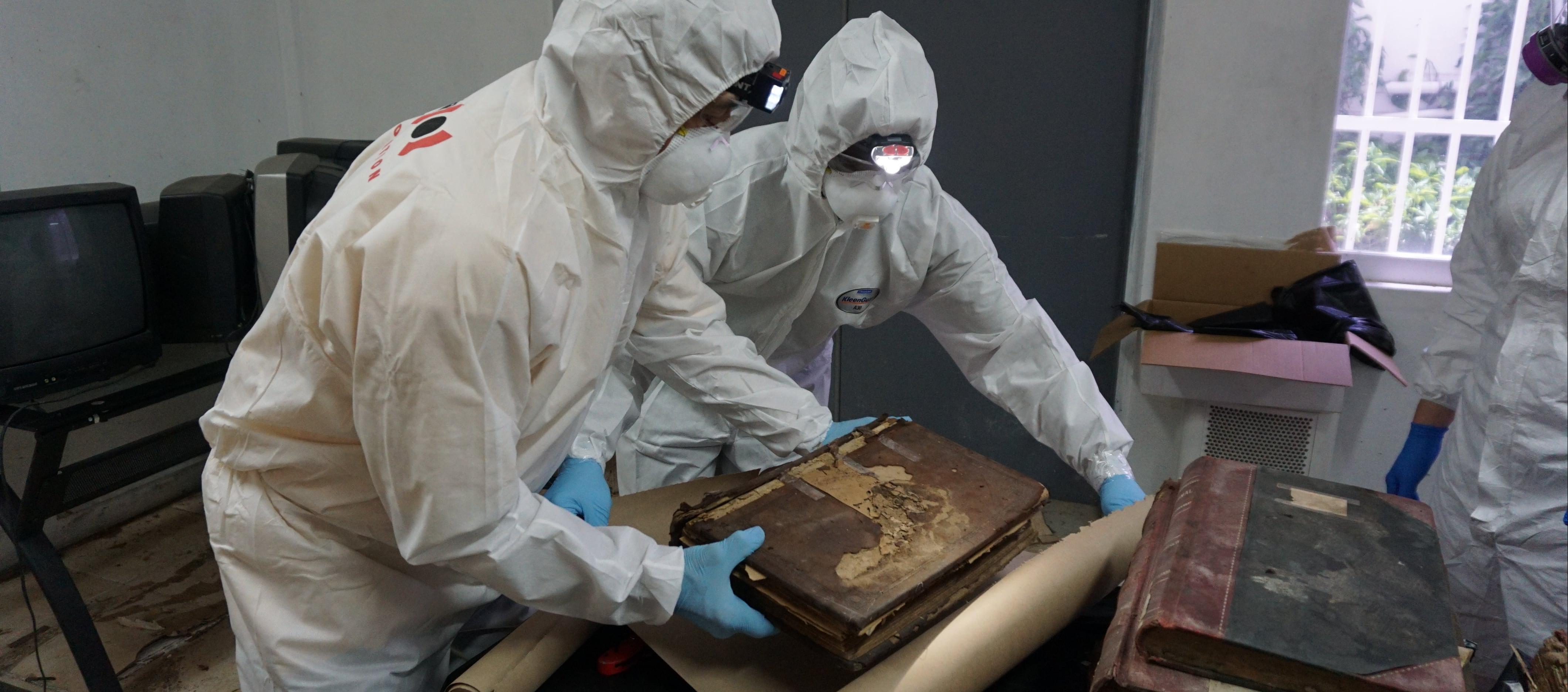 Two people in white hazmat suits handle a damaged historic tome.