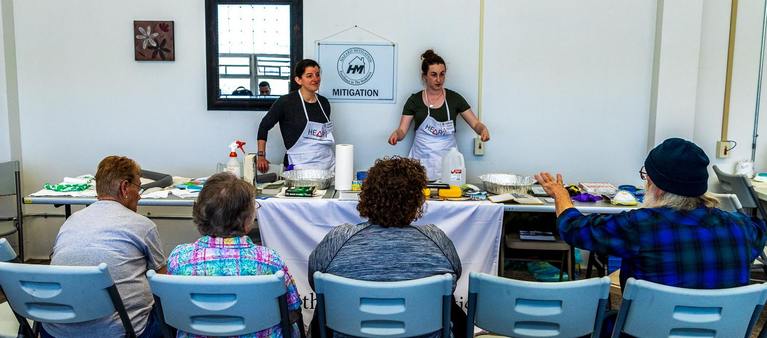 Two women wearing white aprons stand behind table covered with objects.  Four people sitting in chairs look on.  Man in blue shirt points towards table.