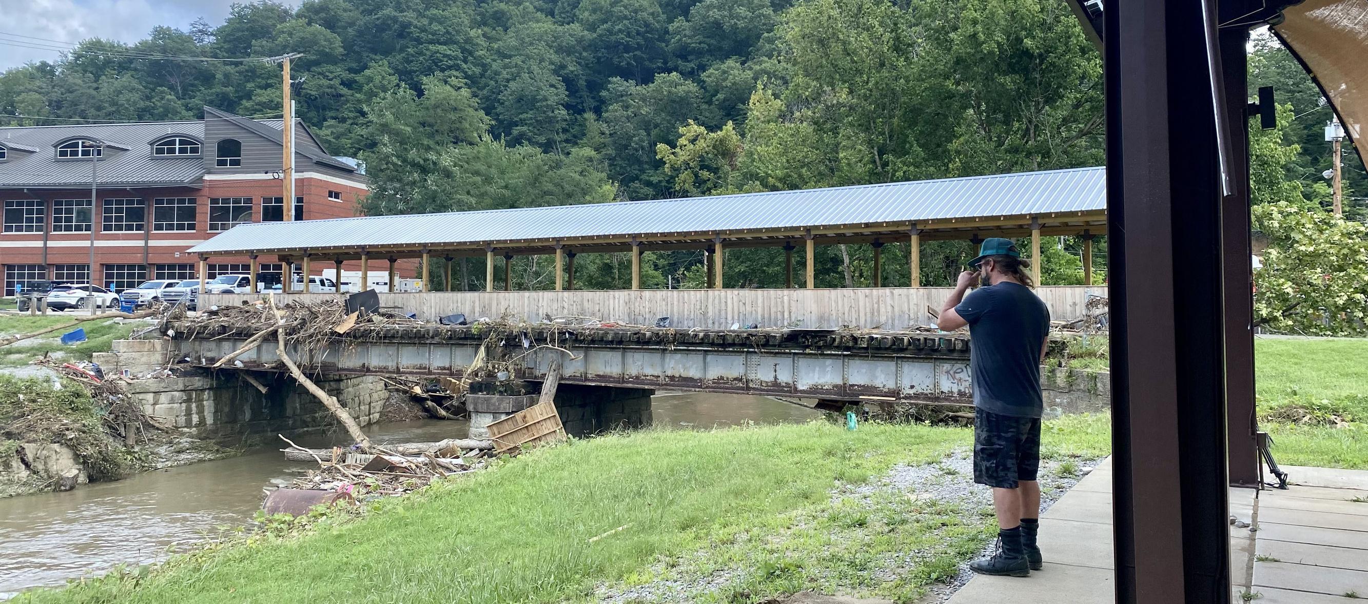 Man wearing black observes debris in river pushing against a covered bridge