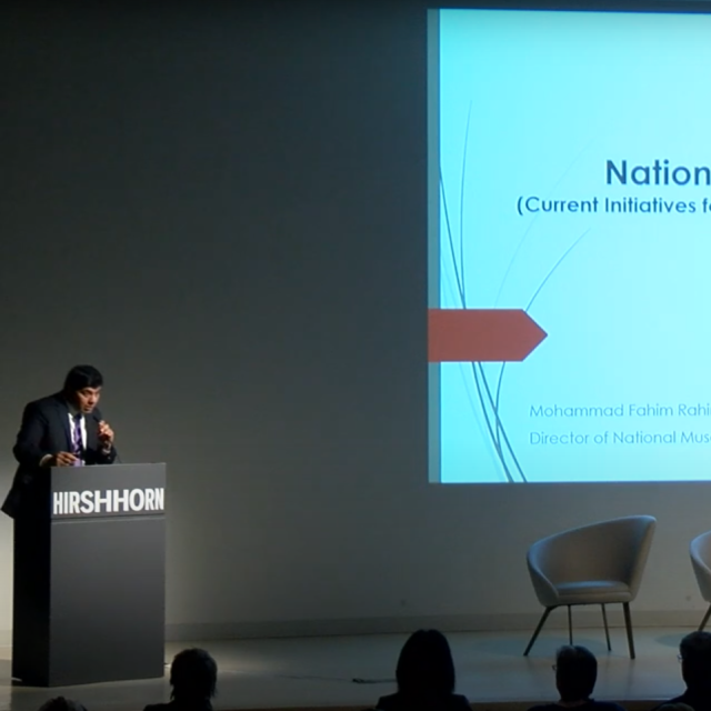 Man in dark gray suit stands at podium. National Museum of Afghanistan on screen