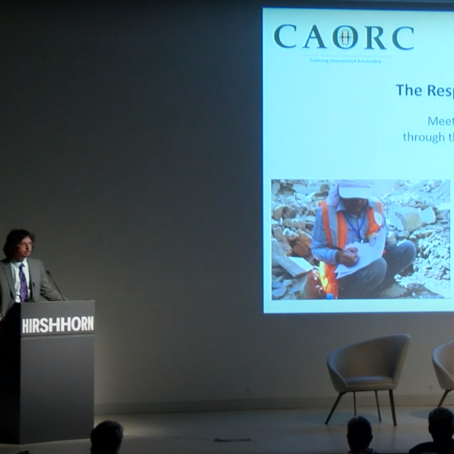 Man in gray suit stands at podium. The Responsive Preservation Initiative on screen