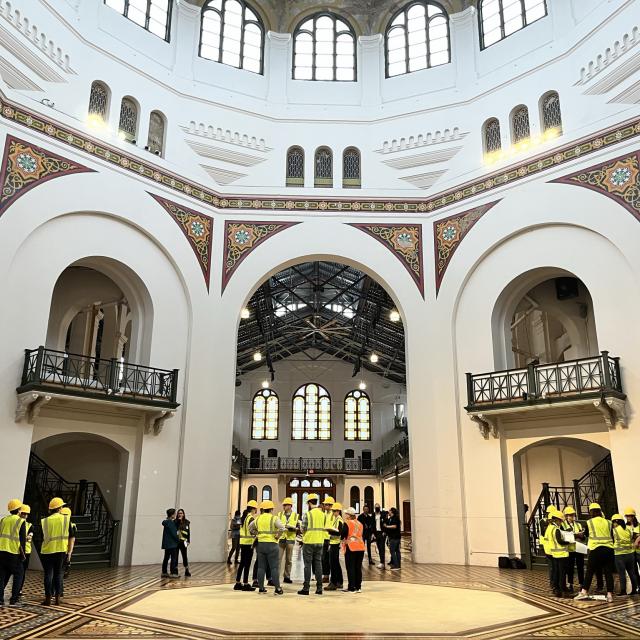 three clusters of people all wearing yellow vests and hardhats stand in the atrium of a building