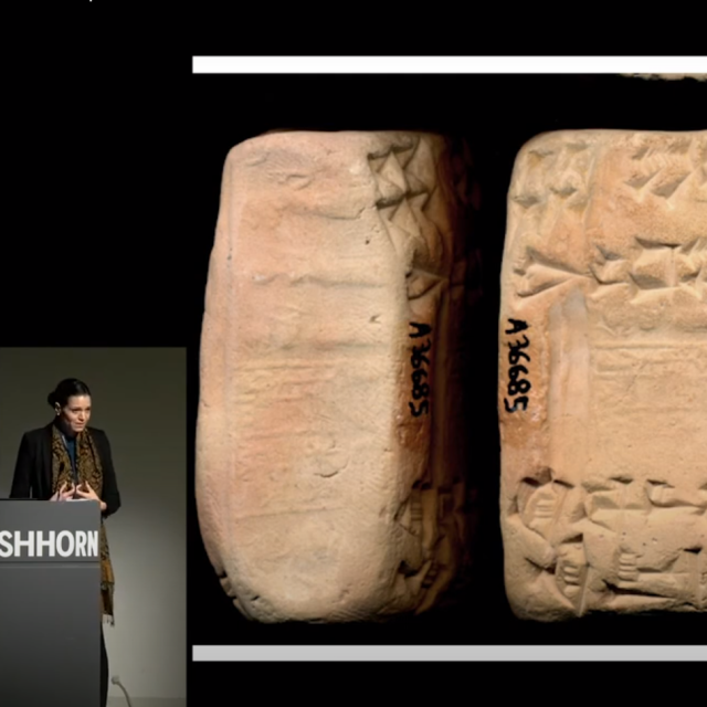 Woman in black jacket and long scarf stands at podium. three (tan squarish in shape) cuneiform tablets on screen