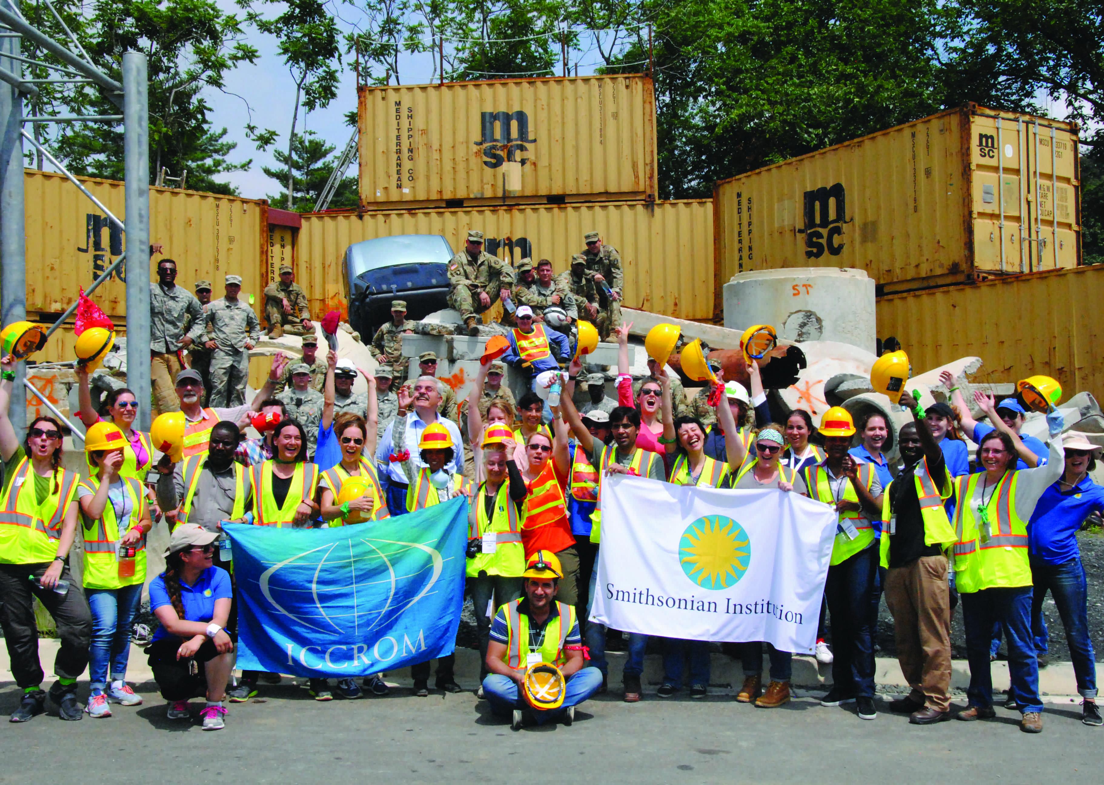 A group of people in yellow vests and hard hats stand in front of shipping containers while holding a blue ICOM flag and white Smithsonian flag