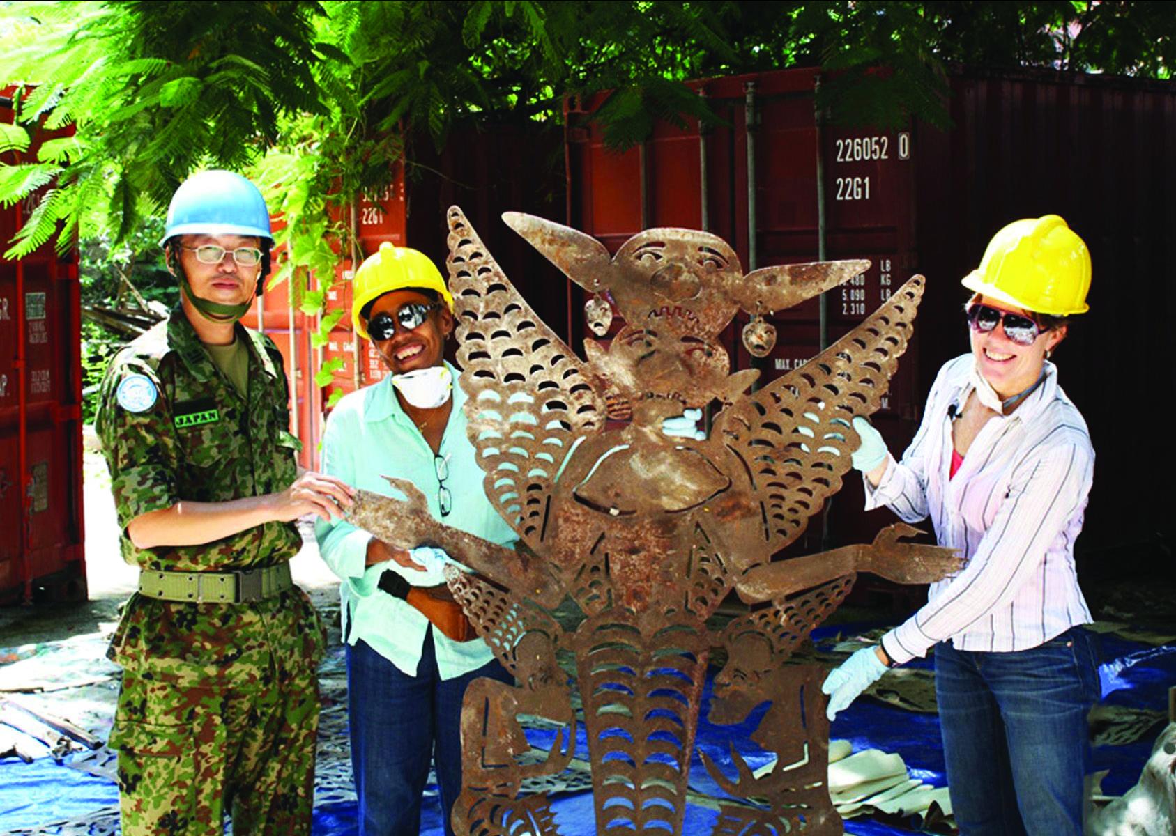 A man in military uniform wearing a blue UN helmet stands with two women in yellow hardhats holding a metal sculpture
