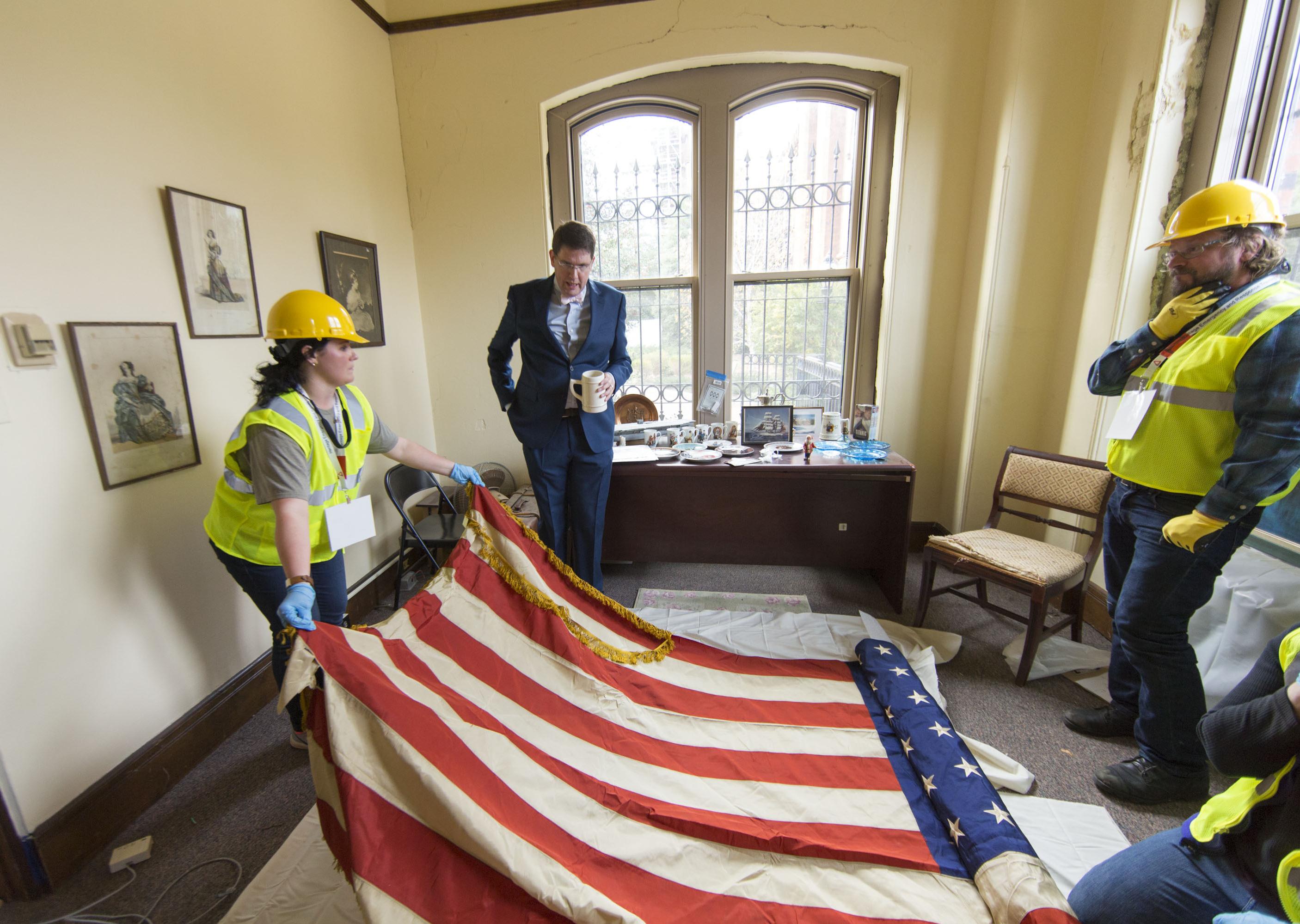 Two Heritage Emergency and Response Training participants in yellow vests and hardhats handle an American flag while a man in a suit looks
