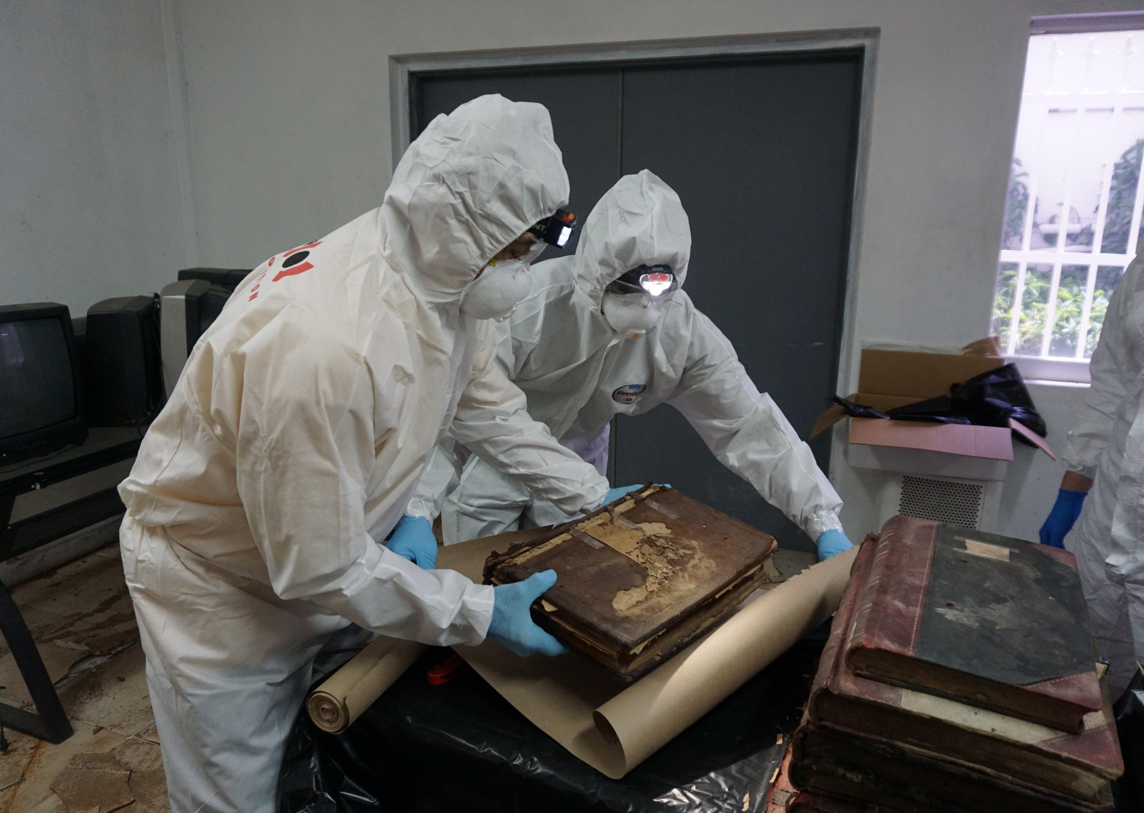 Two people in white hazmat suits handle a damaged historic tome.